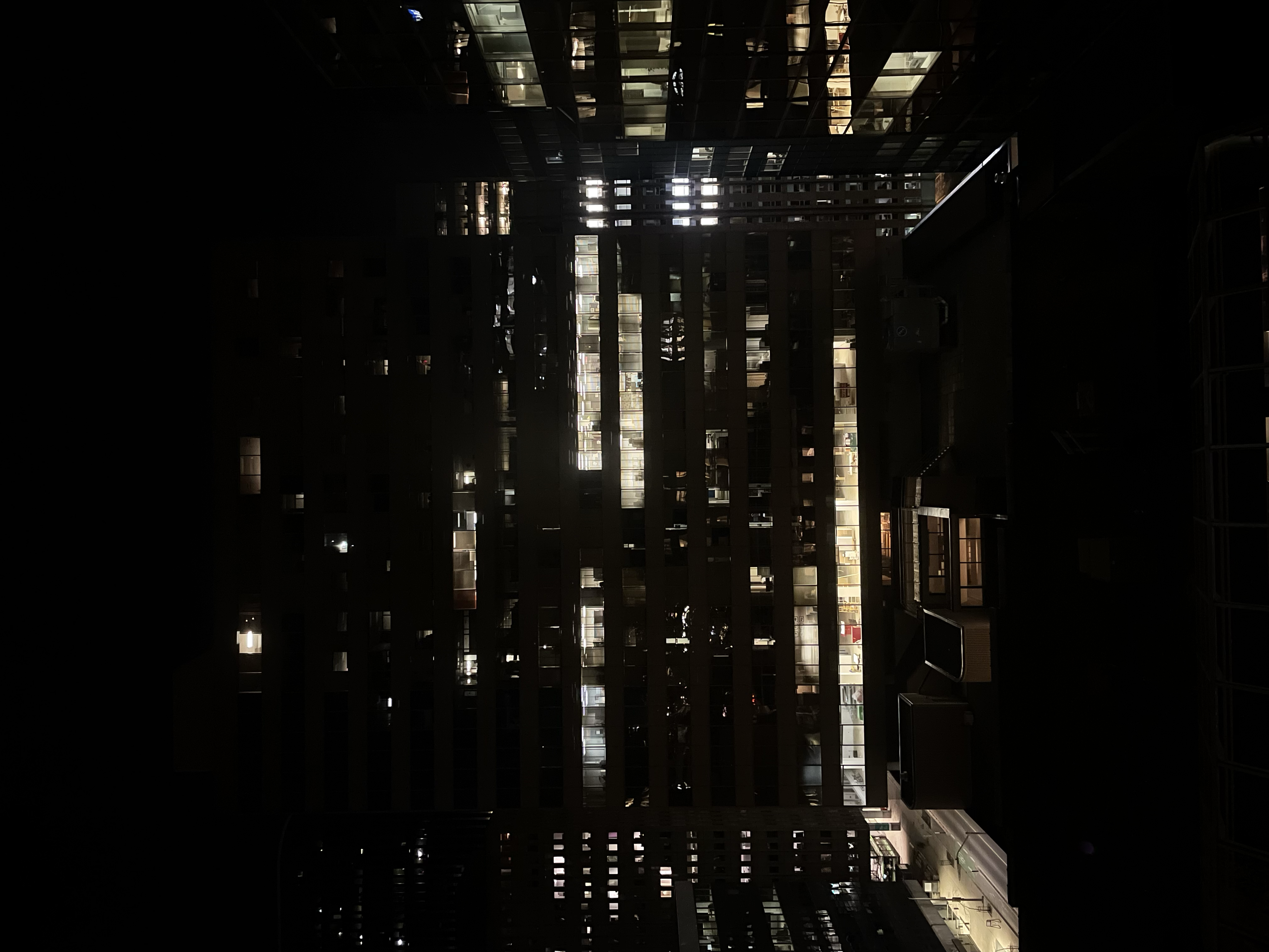 Nighttime falls on a quiet rooftop. The sky is black. In nearby buildings, lit floors reveal open office spaces. Windows glow in the dark.